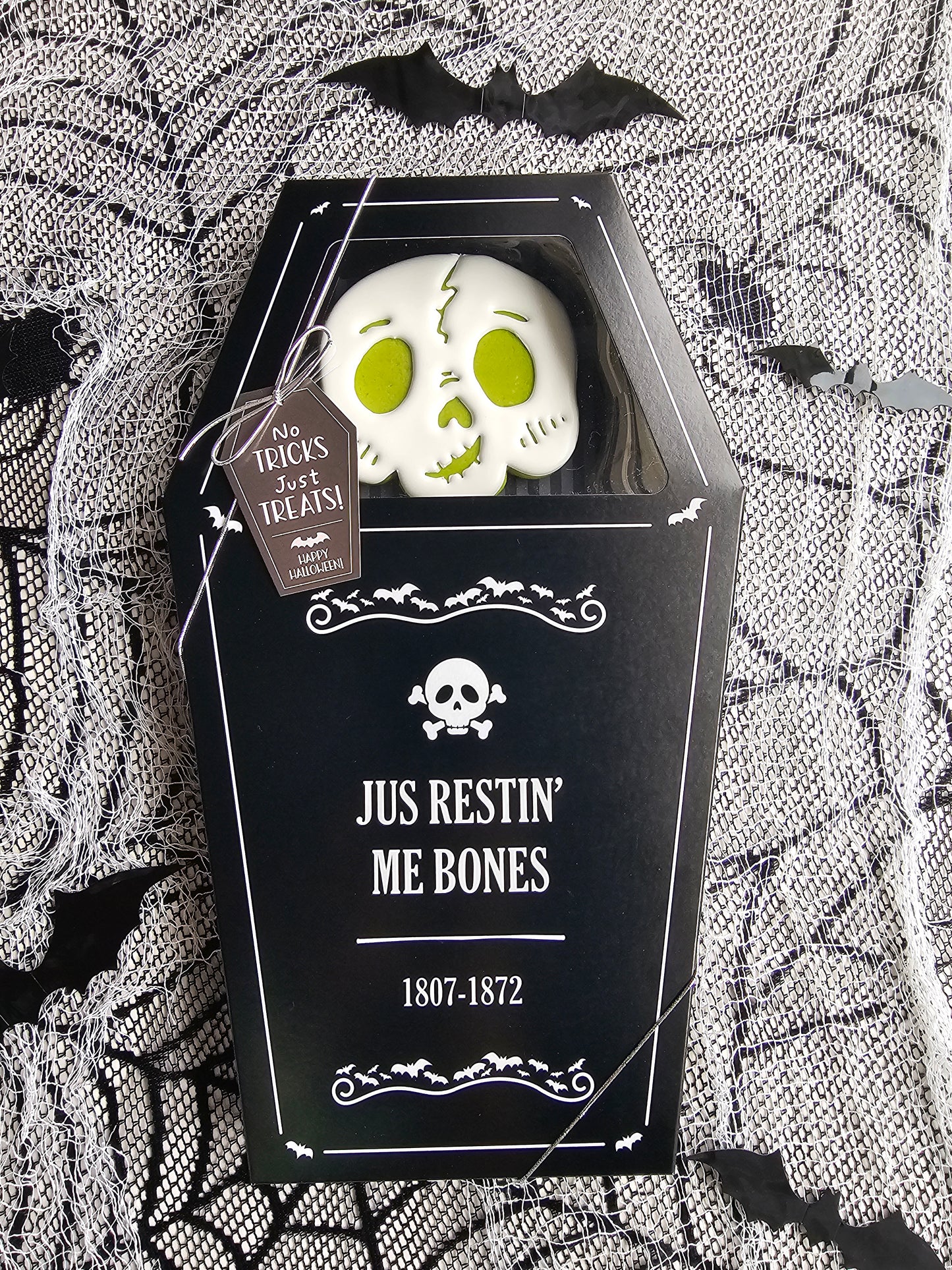 Coffin-shaped black gift box with a skull cookie peeking through a window, Halloween-themed packaging that reads “Jus Restin’ Me Bones.”