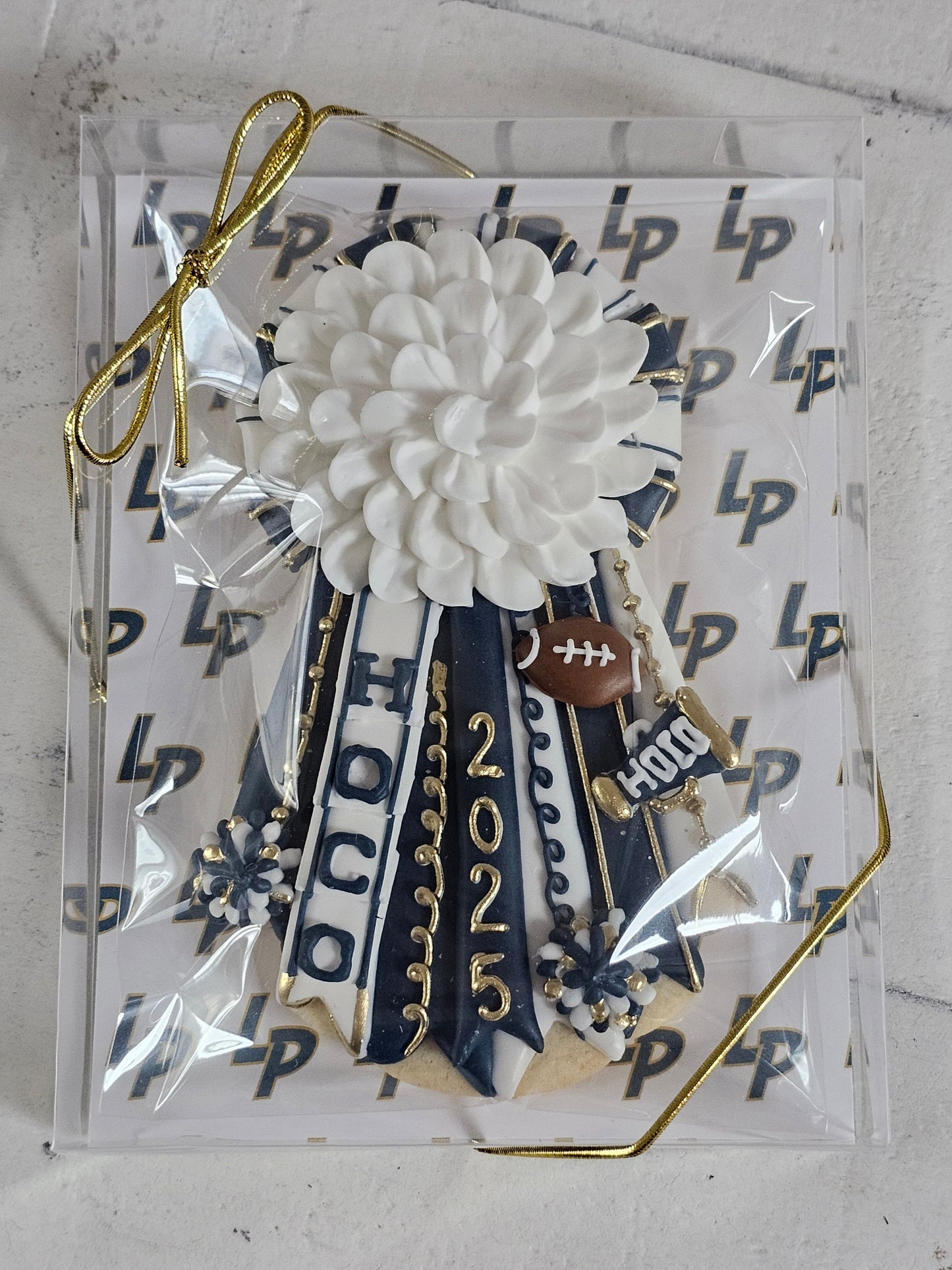 Homecoming mum cookie in white, navy, and gold with Logos Prep School logo background.
