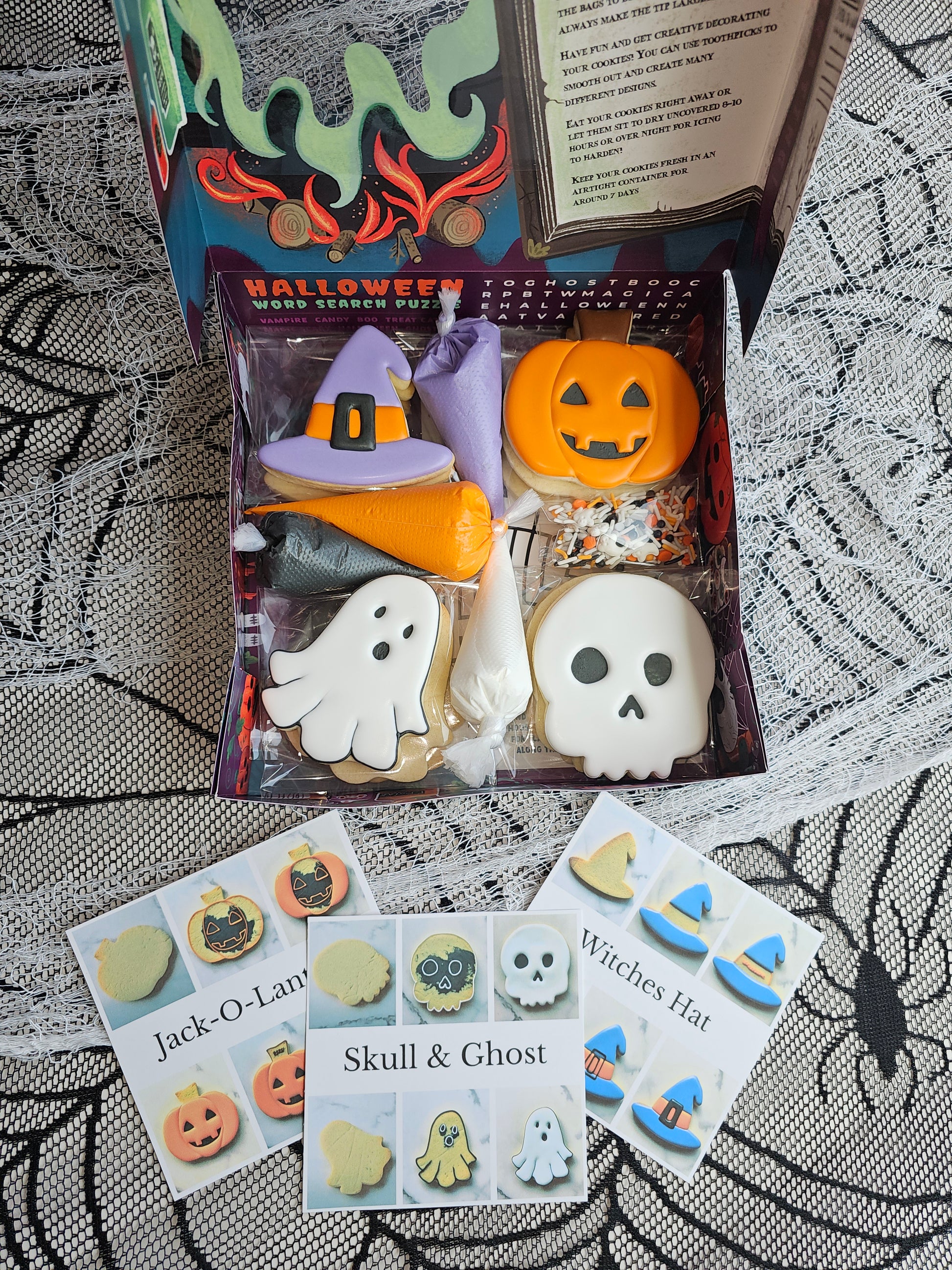 Open Halloween DIY cookie kit in activity box with ghost & witch-hat cookies, four icing bags (orange, black, white, purple) and sprinkles.