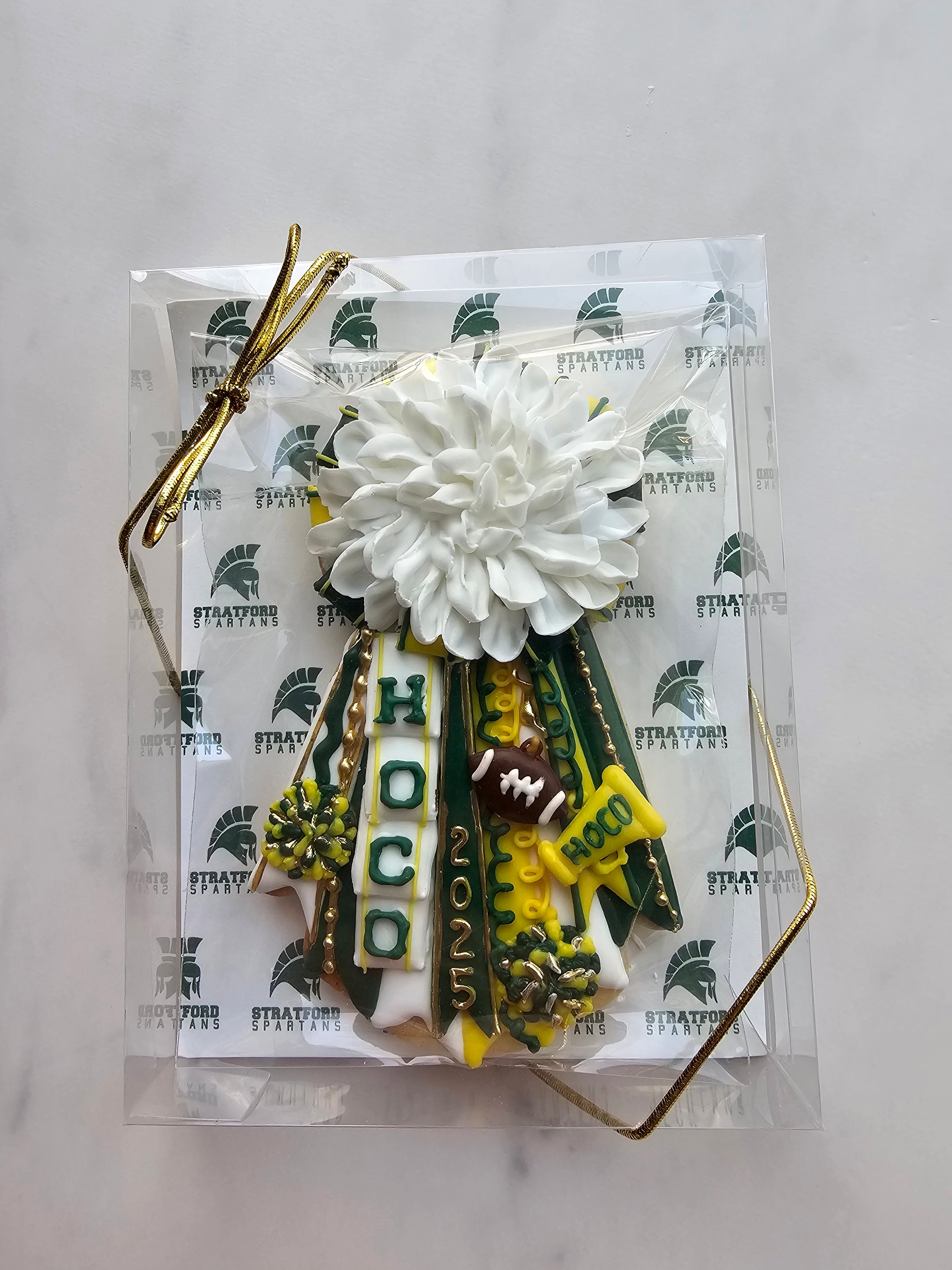 Green, gold, and white homecoming mum cookie with large white chrysanthemum, ‘HOCO’ and ‘2025’ ribbons, football, megaphone, and pom-poms.
